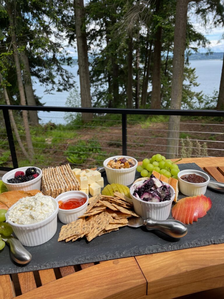 Charcuterie board with crackers, fruit, and cheese on slate with trees in background