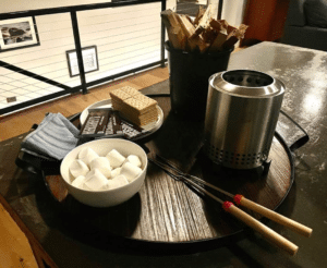 Round wooden tray with round white bowl containing marshmallows, two long forks, two blue striped napkins, a small Solo stove, a bucket filled with wood strips, and a round white plate with Hershey bars and graham crackers