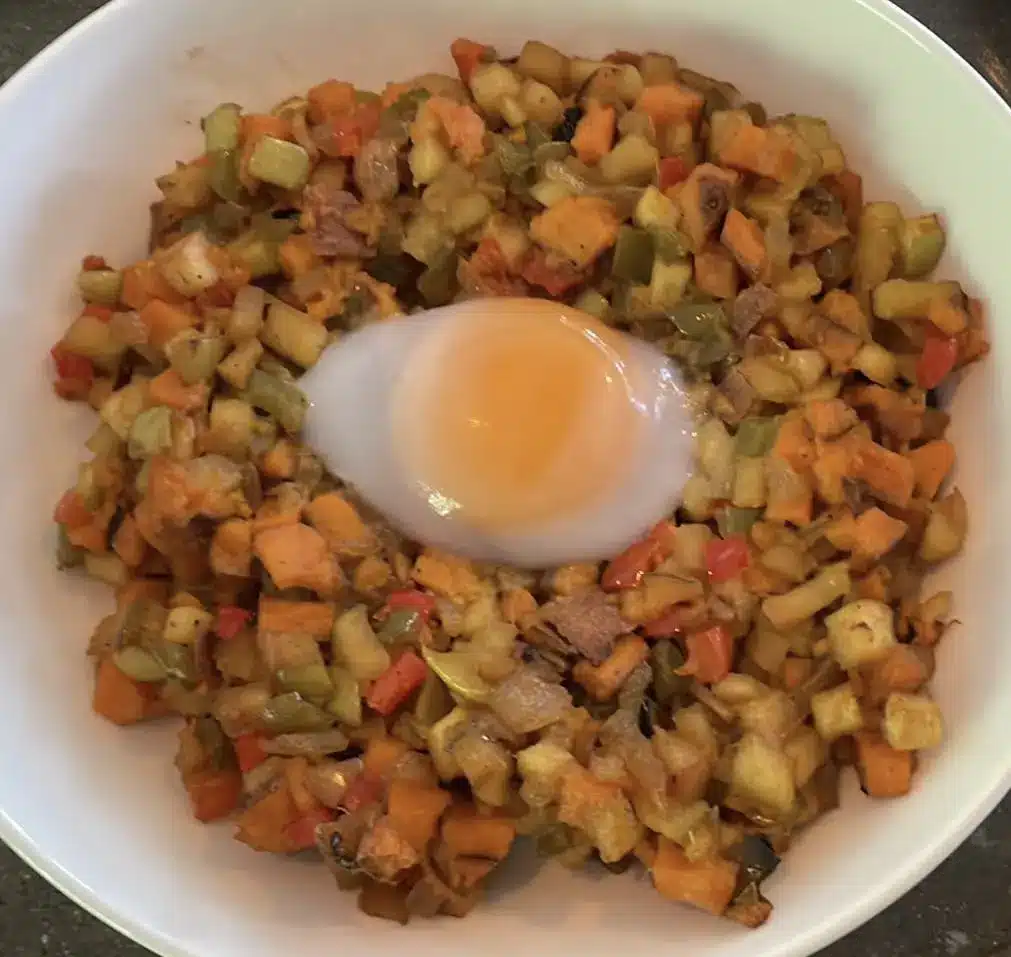 Close-up of colorful bowl of vegetarian hash with onion, sweet potato, apples, and peppers in a white bowl with an egg in the middle